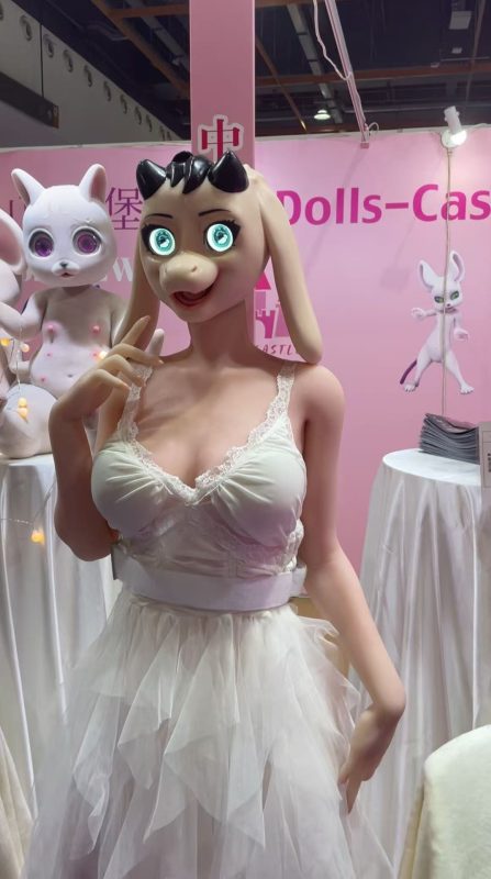 Realistic doll with a cartoonish pig face mask, bright blue eyes, and black hair styled with spikes, wearing a white lace-trimmed dress, displayed at an exhibition with pink background and doll displays.