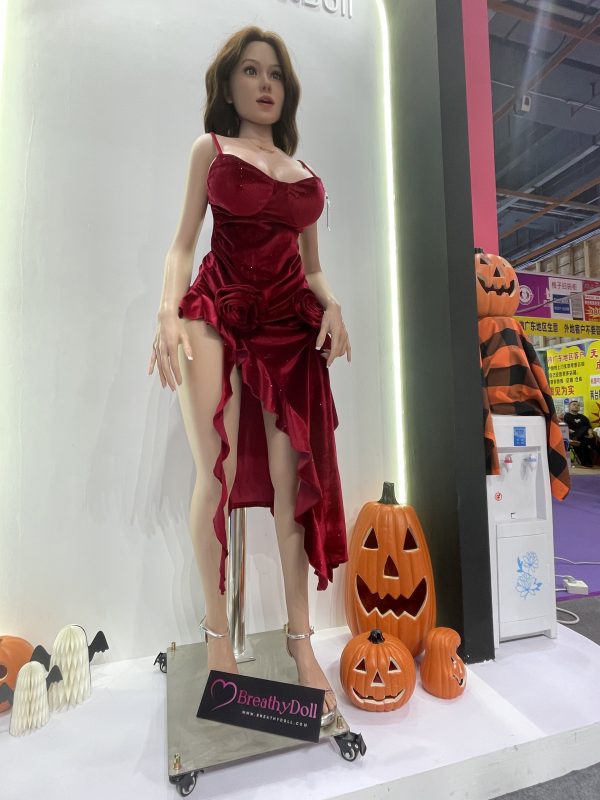 Realistic female doll with long brown hair, dressed in a red velvet dress with floral details, high heels, and lifelike facial features, displayed on a stand with Halloween pumpkins around.