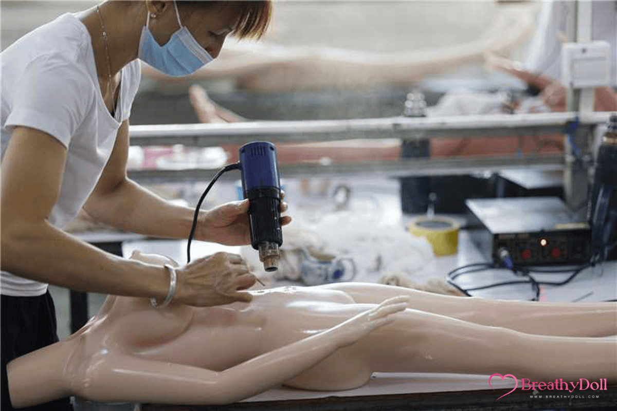 Life-size silicone doll being crafted in a factory, with a worker wearing a face mask using a hot tool to refine the doll's body features, showcasing high-quality manufacturing.