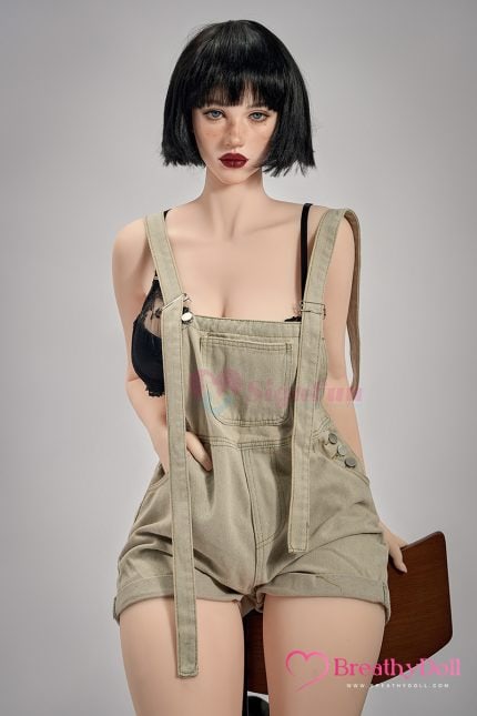 Highly realistic female doll with short black hair, wearing a black lace top and beige overalls, posed sitting on a chair against a neutral background.