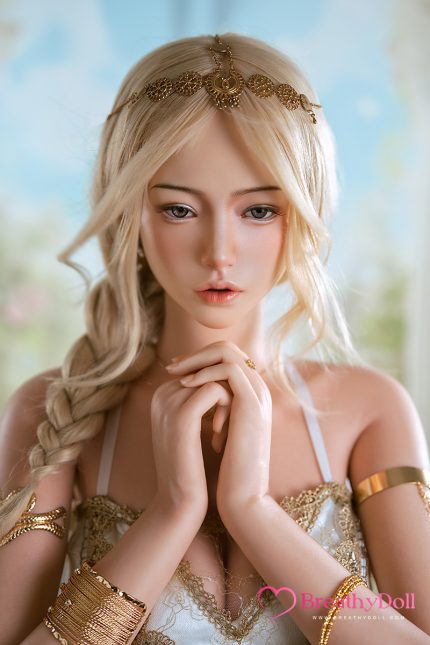 A woman with long blonde hair styled in loose waves, wearing a gold headpiece and elegant jewelry, gazing softly with her hands near her face against a blurred outdoor background.