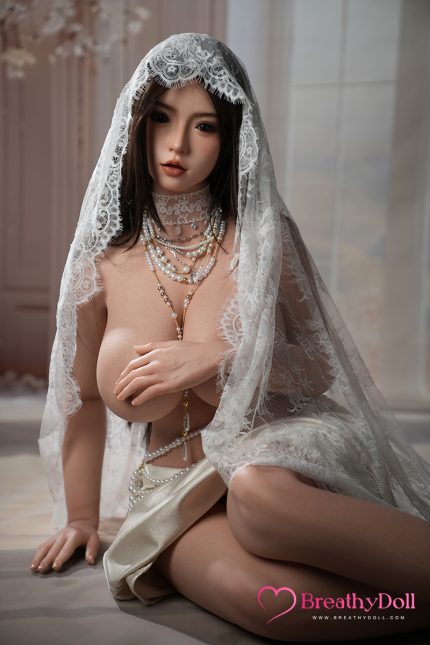 A woman with long dark hair, wearing a lace veil and multiple pearl necklaces, sitting on the floor with a sheer fabric draped over her shoulders in a softly lit room.