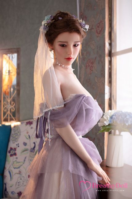 Young woman in a lavender dress with floral headpiece and veil, standing indoors near a window with floral wallpaper.