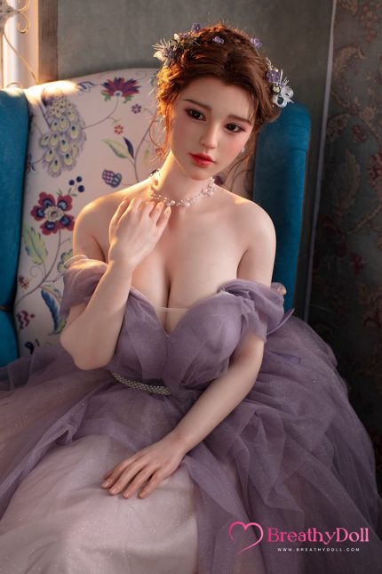 Realistic female doll with brown hair styled in an updo, wearing a purple gown and floral hair accessories, sitting on a blue armchair in a decorated room.
