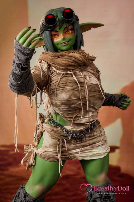 Cosplay of a goblin character with green skin, black hair, and goggles, wearing a tattered beige outfit, standing indoors.